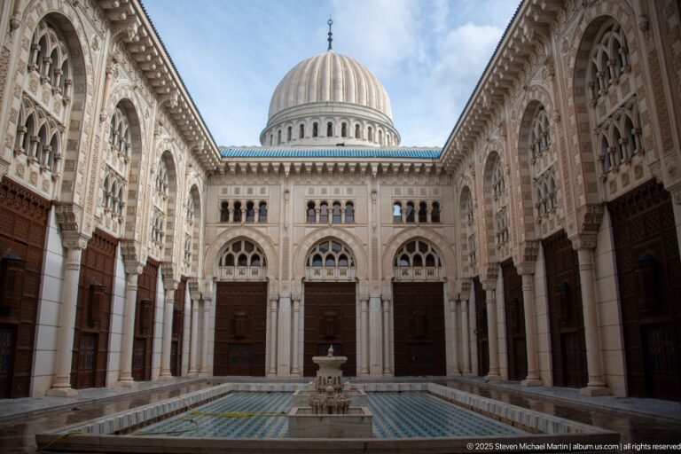 Pale arcaded mosque courtyard with a small central fountain, tall wooden doors, carved openings, and a ribbed dome beyond.