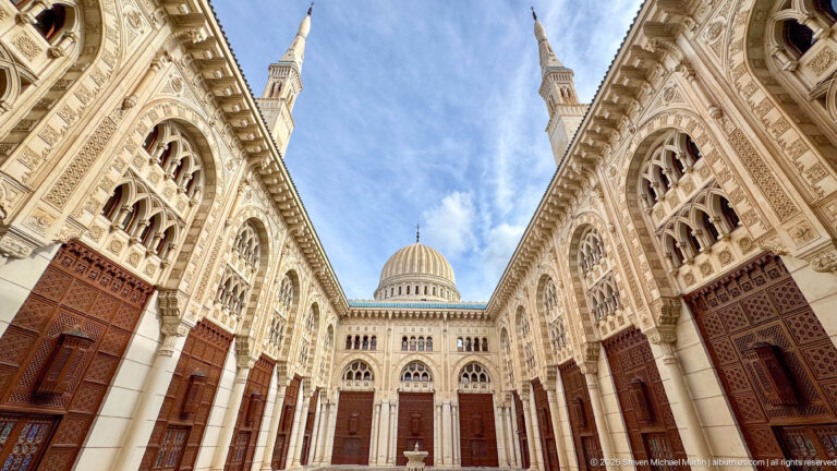 Upward view through an ornate mosque courtyard toward a ribbed dome, blue sky, and two slender minaret tips.