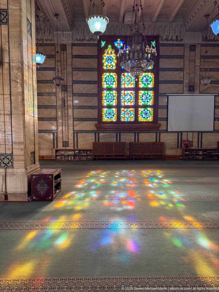 Wide spread of multicolored stained-glass reflections across green prayer carpet, with the bright window and chandelier beyond.