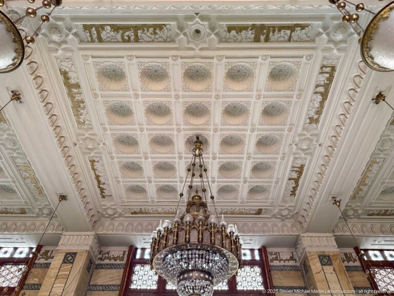 Large crystal chandelier hanging beneath a pale coffered ceiling decorated with rosettes, relief ornament, and gilded accents.