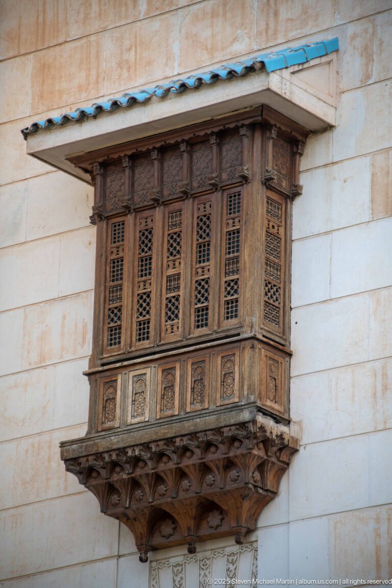 Front view of a dark carved wooden projecting window with lattice shutters on a pale stone façade.