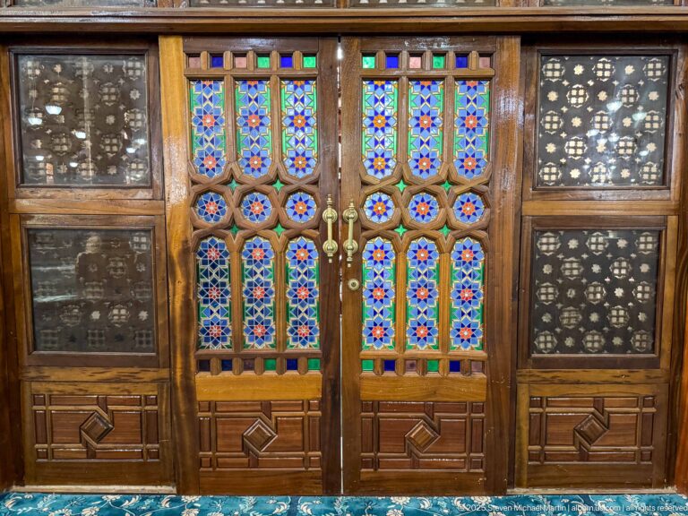 Wooden double doors with multicolored glass inserts, carved lower panels, and dark pierced geometric screens on both sides.