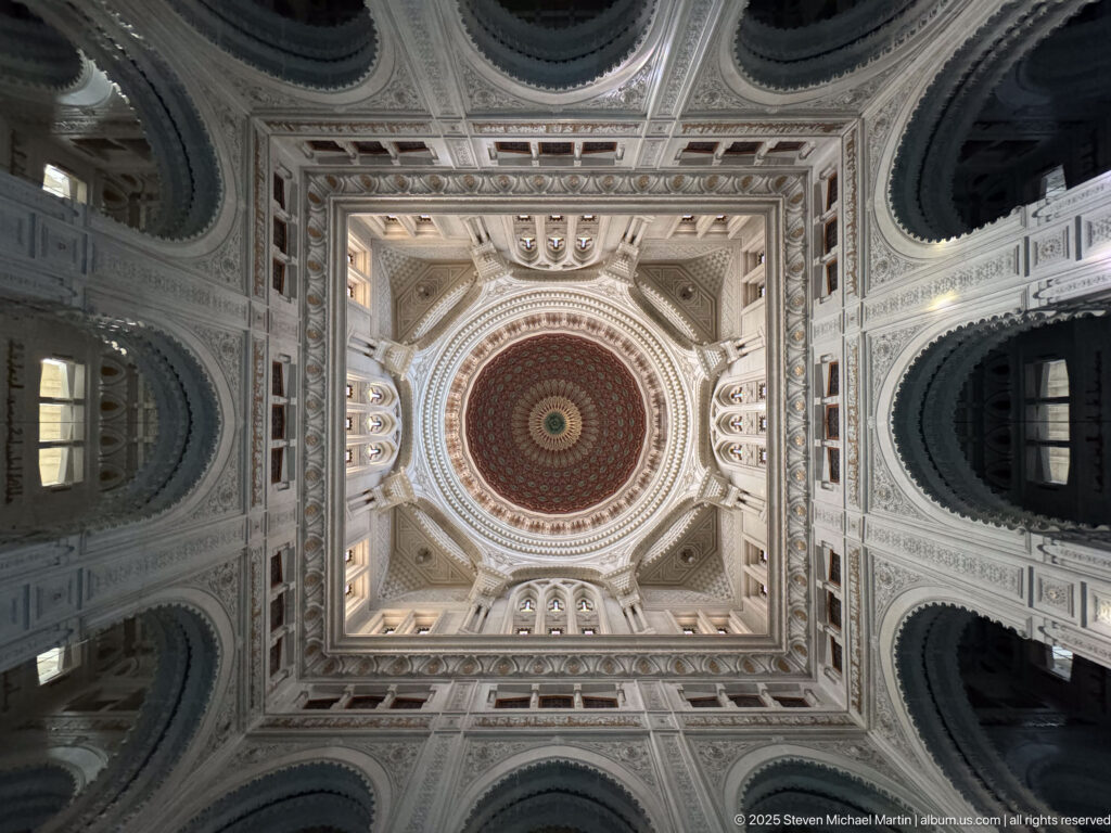 Upward symmetrical view of a richly decorated circular dome centered within pale arches, recessed bays, and square structural framing.