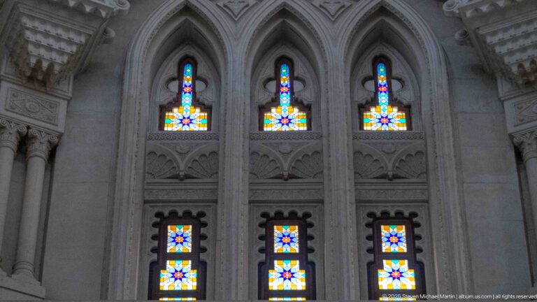 Six colorful geometric stained-glass windows arranged in two tiers within pointed arches and carved pale wall decoration.