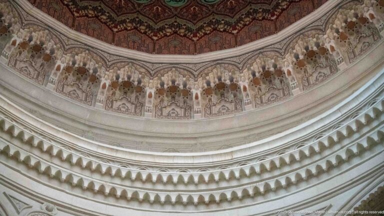 Close view beneath a dome showing tiered niche-like relief ornament below a red, gold, and green painted ceiling surface.