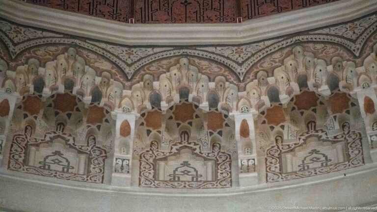 Tiered muqarnas-like niches and painted geometric details beneath a curved band of Arabic calligraphy.