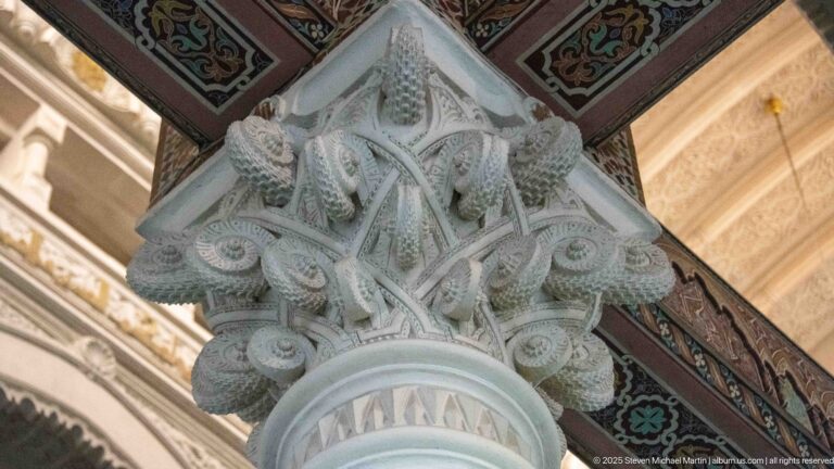 Close upward view of a pale carved column capital beneath a beam decorated in red, green, black, and gold patterns.