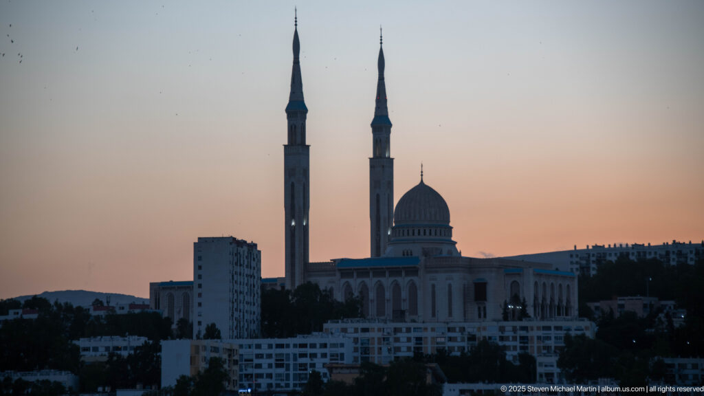 Distant dusk view of Amir Abdel Kader Mosque with twin slender minarets and a central dome silhouetted above city buildings.