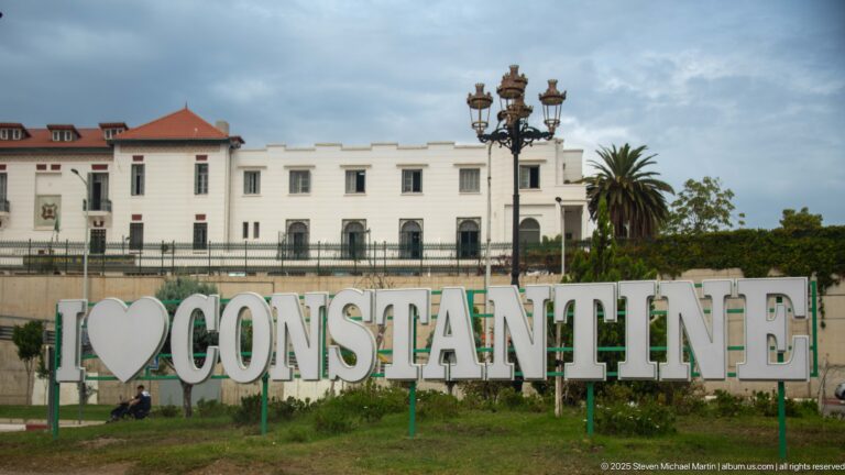 Large “I love Constantine” sign in a landscaped public space with white buildings, palms, and ornate streetlights behind it.