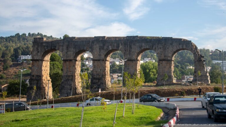 Weathered stone aqueduct arcade with tall semicircular arches beside a modern road, cars, grass, and young trees.