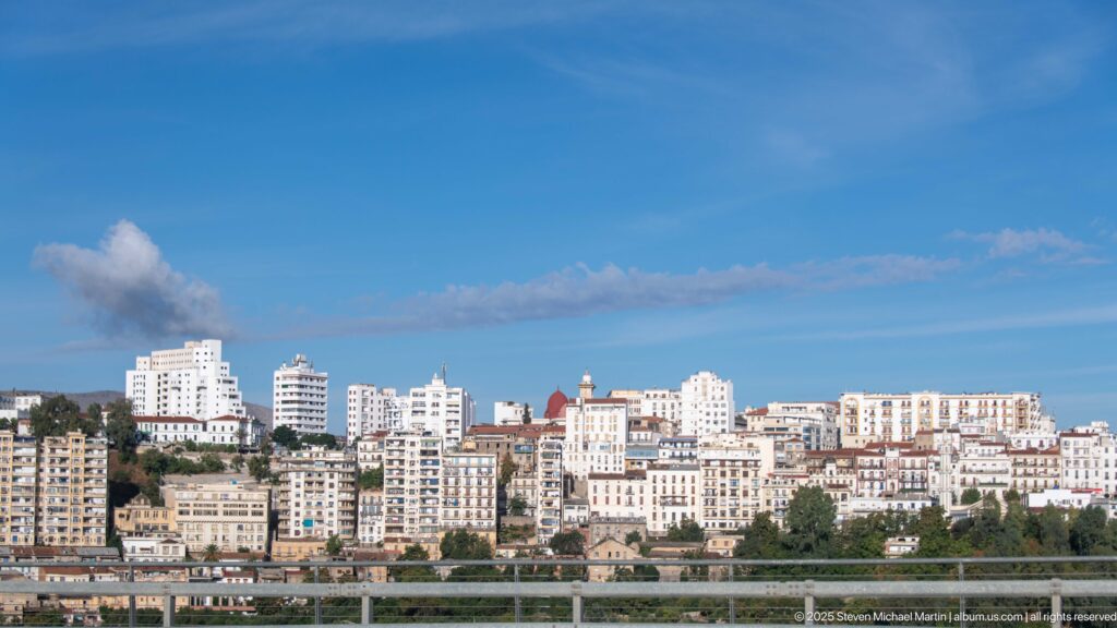 Hillside skyline of Constantine with dense white apartment buildings above an elevated roadway beneath a clear blue sky.