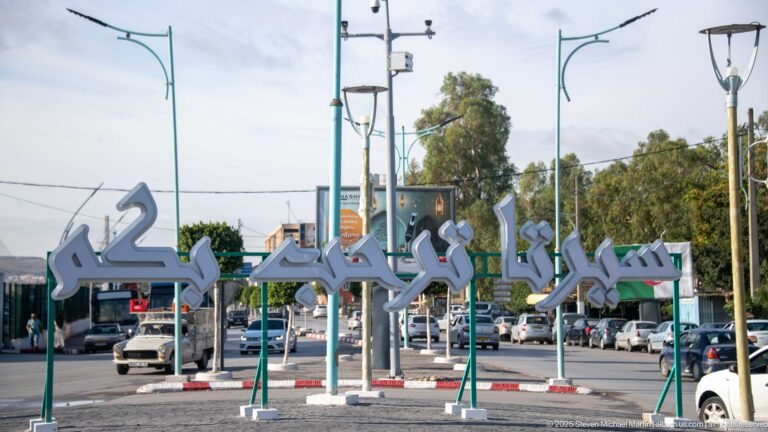 Large freestanding Arabic-script sign beside a busy Constantine road with cars, streetlights, and roadside landscaping.