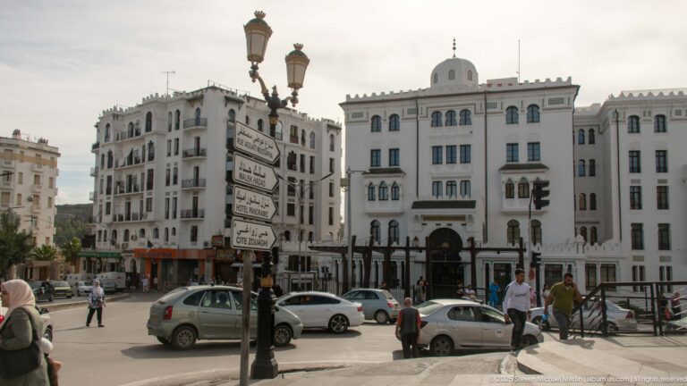 White multi-story civic buildings around a Constantine intersection with cars, pedestrians, street lamps, and Arabic-French direction signs.