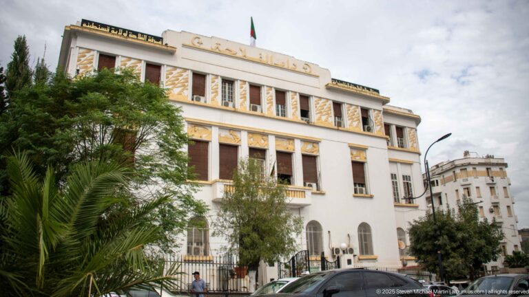 White civic building with tall windows, gold decorative panels, Arabic roofline lettering, an Algerian flag, palms, and street trees.