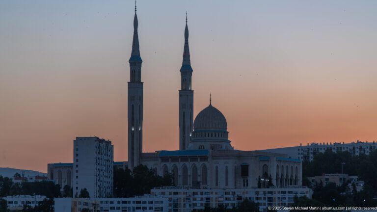 Emir Abdelkader Mosque at dusk, with a central dome and two tall slender minarets silhouetted against a peach-blue sky.