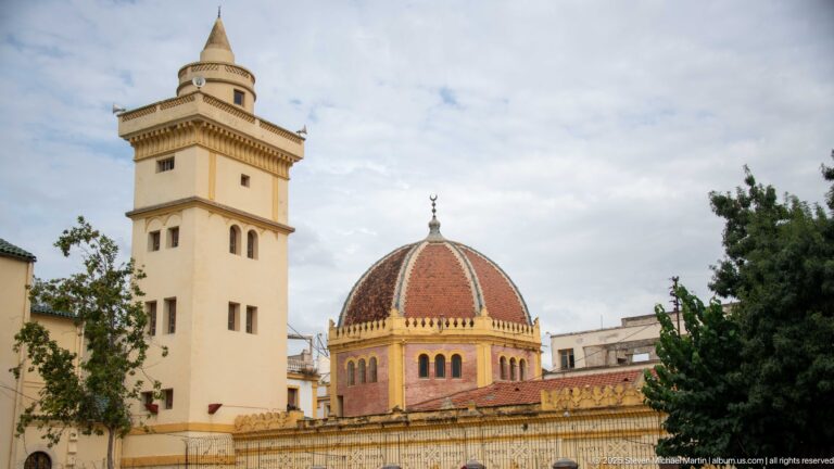 Square cream-colored tower beside a red-and-dark patterned dome topped with a crescent, partly screened by trees and buildings.