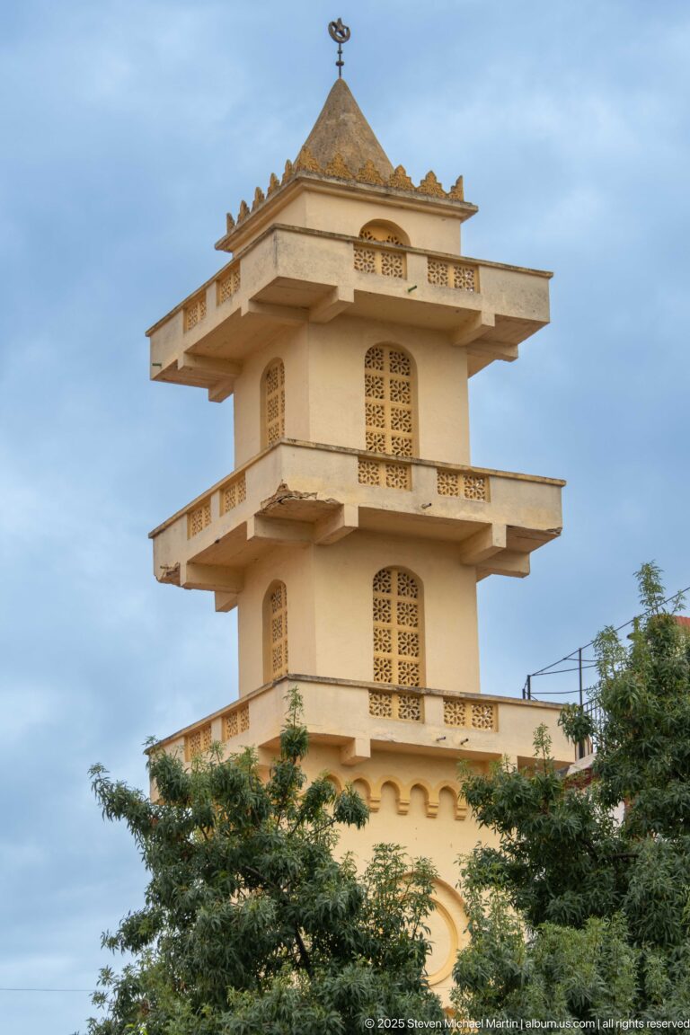 Pale tiered square tower with broad projecting eaves, perforated geometric screens, arched openings, and a small finial.