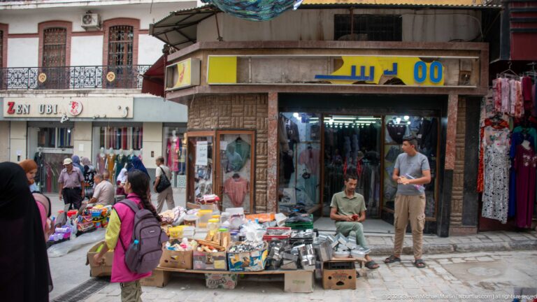 Sidewalk vendor selling household goods outside narrow Constantine shopfronts as pedestrians pass through the busy commercial street.