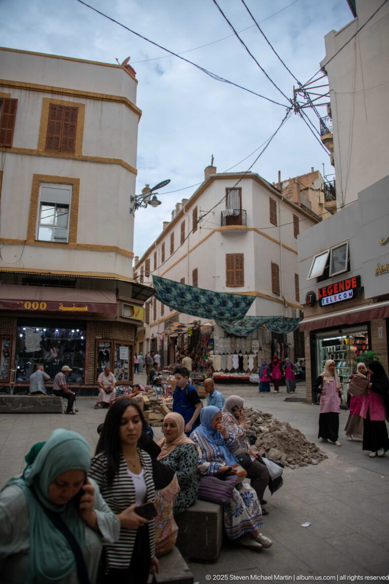 Crowded Constantine market street with shoppers, seated women, storefronts, awnings, and overhead utility wires.