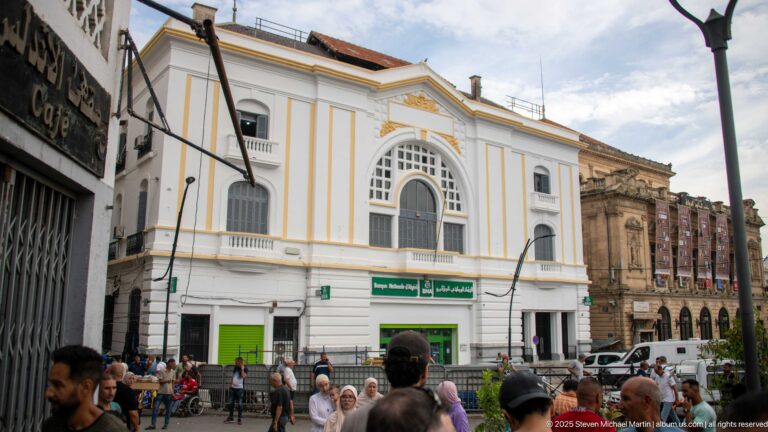 White Banque Nationale d’Algérie building with gold trim, a tall arched window, green bank signs, and crowds at street level.
