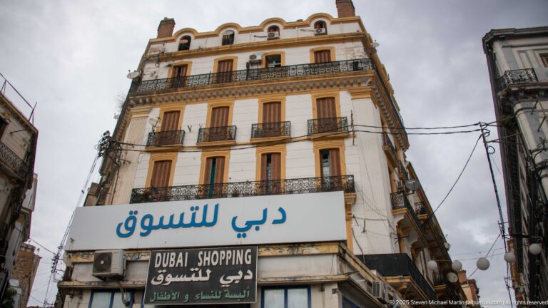 Tall apartment building above a Dubai Shopping storefront, with Arabic signage, wrought-iron balconies, shutters, and overhead cables.