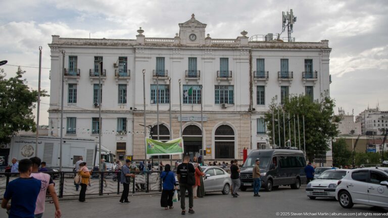 Large white civic building with three entrance arches, tall windows, balconies, and a busy square with pedestrians and vehicles.