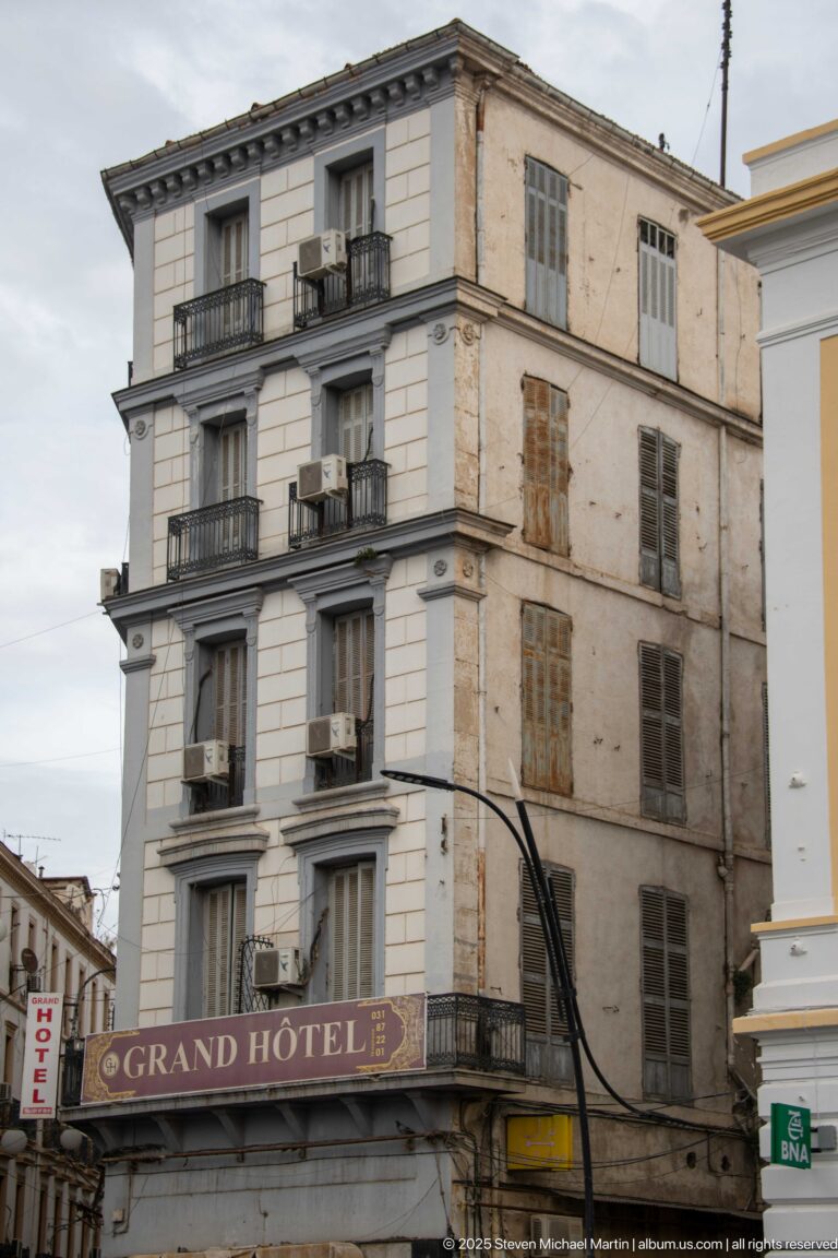Weathered Grand Hôtel corner façade with stacked wrought-iron balconies, shuttered windows, air-conditioning units, and a large hotel sign.