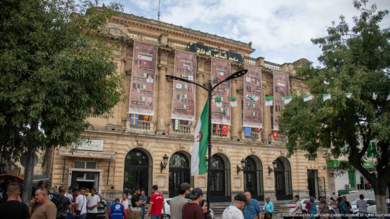 Crowd outside a monumental stone building with tall arched openings, vertical banners, Algerian flags, and trees.