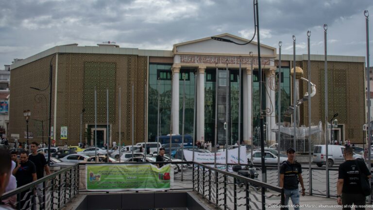 Modern public building with a central glass entrance, geometric screen walls, flagpoles, plaza railings, people, and parked cars.