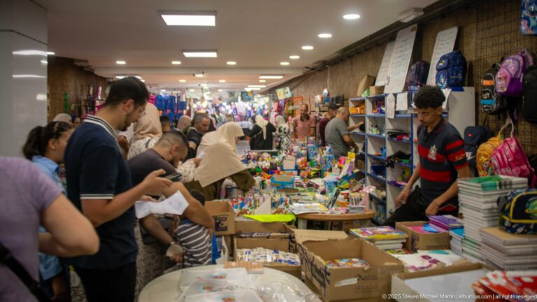 Crowded indoor market aisle with shoppers browsing notebooks, backpacks, toys, and school supplies at densely packed stalls.