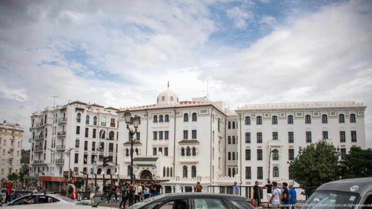 Large white building complex with arcades, tall windows, a small central dome, pedestrians, and traffic on a Constantine square.