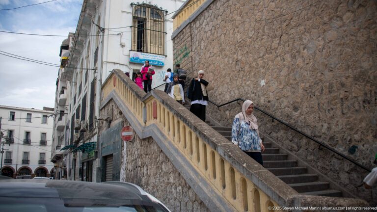 Pedestrians on a steep stone stairway beside a rough masonry wall and closely spaced urban buildings.
