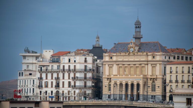 Historic civic building with arched façade, central clock, patterned roof, slender tower, and apartment blocks alongside.