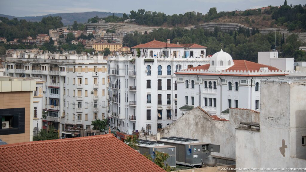 Elevated view across white mid-rise buildings with red tile roofs and a small dome, backed by green Constantine hillsides.