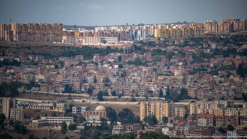 Telephoto panorama of dense Constantine neighborhoods climbing hillsides, with apartment blocks on the ridge and a dome in the middle distance.