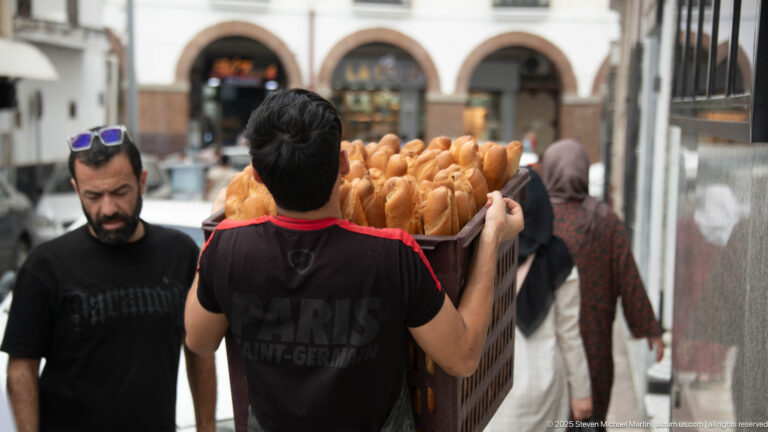 Worker carrying a large shoulder-high tray packed with baguettes through a busy market passage in Constantine.