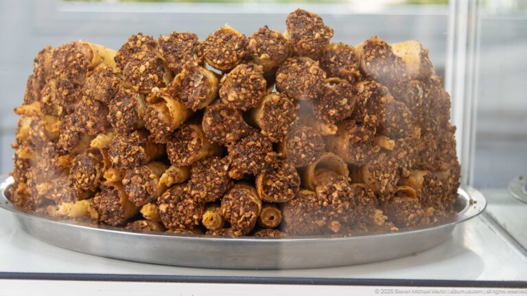 Close-up of rolled brown pastries heavily coated with sesame seeds and syrup, piled on a metal tray behind glass.