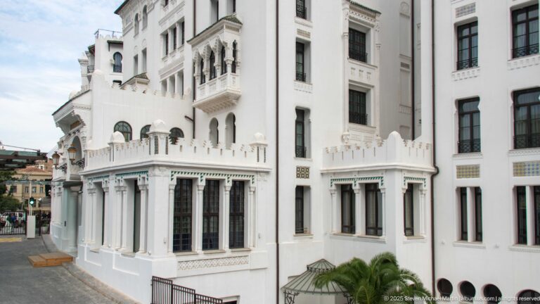 White hotel façade with projecting bays, balconies, scalloped arches, pierced details, green-tiled canopies, and dark windows.