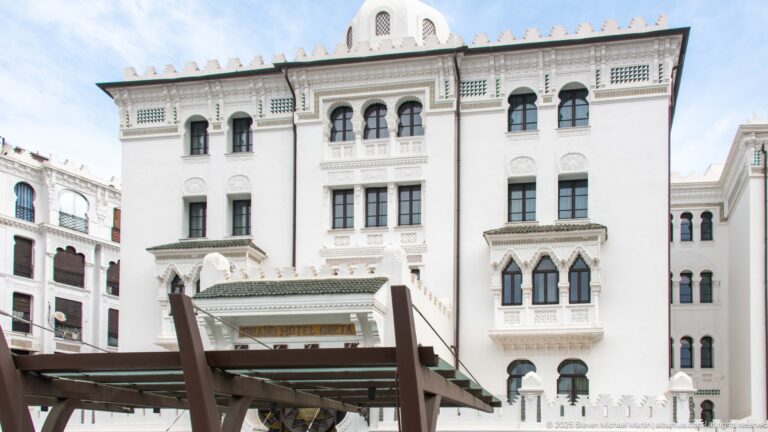 Tall white hotel entrance façade with arched windows, decorative balconies, green-tiled awnings, carved panels, and a timber entrance canopy.