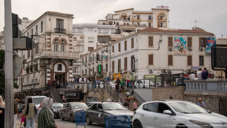 Busy Constantine street with cars, pedestrians, shops, steps, balconies, and closely packed white buildings.