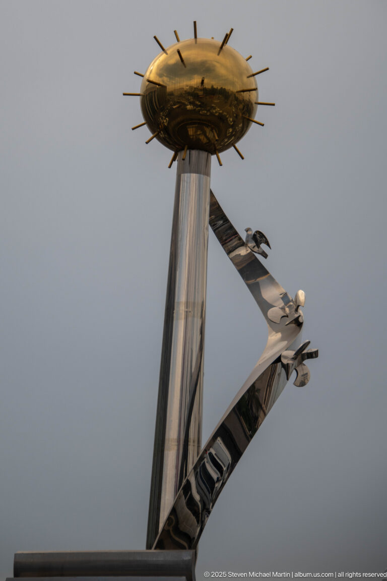 Modern Constantine monument with tall metallic column, gold circular crown, and diagonal sculptural element against the sky.