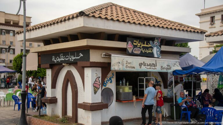 Tiled-roof ice-cream and sweets kiosk with Arabic signage, customers, blue chairs, and market tents.