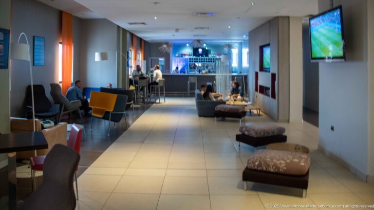Contemporary hotel lounge with tiled floor, chairs, benches, televisions, and a bar at the far end.