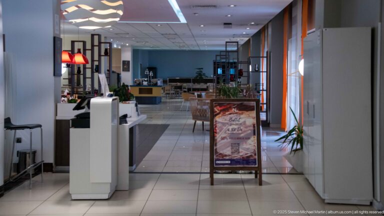 Bright hotel lobby corridor with reception counters, lounge furniture, plants, and orange window accents.