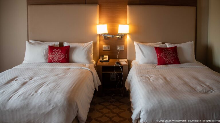 Two twin hotel beds with white linens, red embroidered pillows, paired wall lamps, and a shared bedside table.