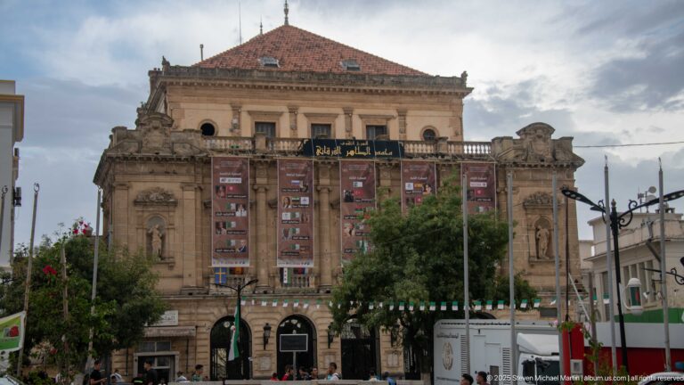 Ornate historic stone civic building with tall windows, sculpted façade, banners, trees, and a tiled roof.