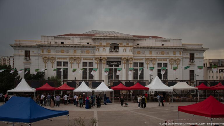 Large symmetrical white civic building behind red and white event tents and people in a Constantine square.