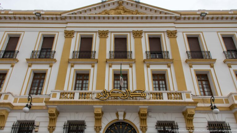 White-and-ochre institutional building with pilasters, balconies, triangular pediment, and gold Arabic lettering above the central entrance.