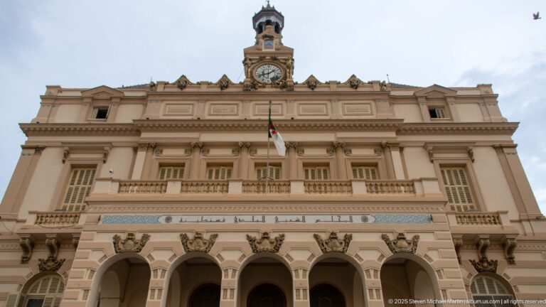 Constantine city hall façade with arched entrance, columns, Algerian flag, Arabic inscription, and central clock tower.