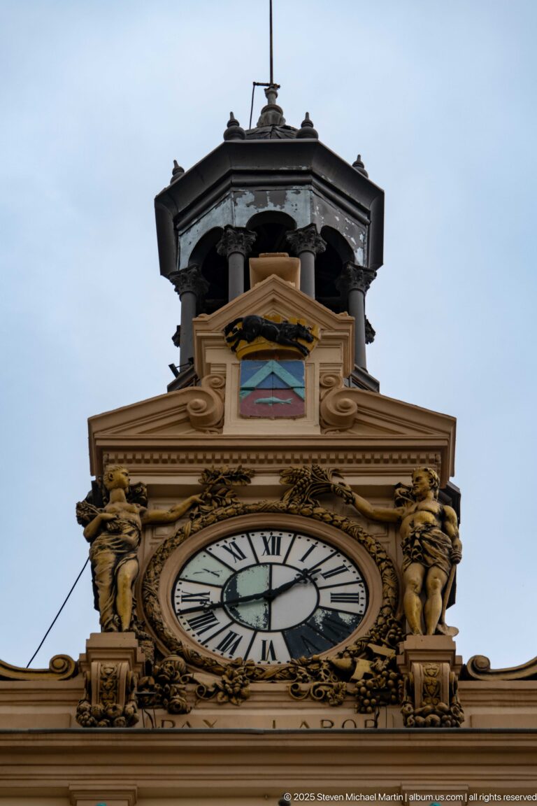 Close view of Constantine city hall clock with Roman numerals, sculpted figures, ornament, and open belfry above.